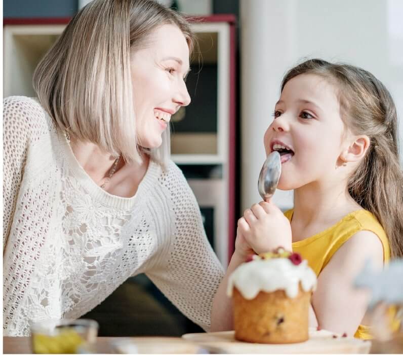 An adult and a child smile as the child licks a spoon. An iced cake is on the table in front of them.
